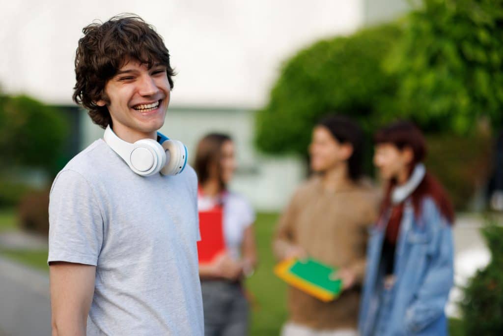 Portrait of a cheerful male student wearing headphones on a university campus with his classmates chatting in the background